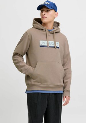 Sweat à capuche marron en tissu doux, doté d'une poche avant et d'une étiquette à impression graphique. Superposé à une chemise bleue et associé à un pantalon foncé.