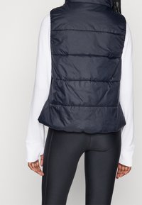 Weste in Marineblau mit horizontalen Steppmustern, einem hohen Kragen und seitlichen Klappen, getragen über einem weißen Langarmoberteil.