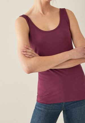 Femme portant un haut sans manches bordeaux et un jean bleu, les bras croisés devant un arrière-plan neutre.