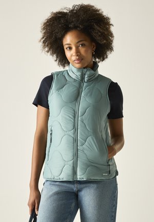 Vrouw met krullend haar die een lichtblauw doorgestikt vest draagt over een zwart T-shirt met korte mouwen en een blauwe spijkerbroek, staande met één hand in een zak.