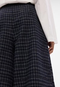 Hlače od vune s kariranim uzorkom u tamnoplavoj boji sivo-smešnim šarama. Imaju opušteni kroj i široke nogavice, u kombinaciji s belom košuljom.