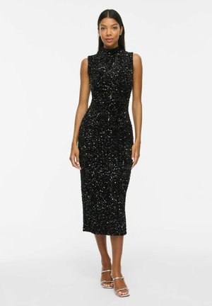 Mujer con vestido midi negro ajustado sin mangas con lentejuelas, cuello alto y tacones plateados con tiras sobre fondo blanco liso.