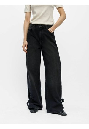 Personne portant un jean noir à jambes larges avec des liens aux ourlets, une chemise beige à manches courtes, des chaussures noires, debout avec une main dans la poche.