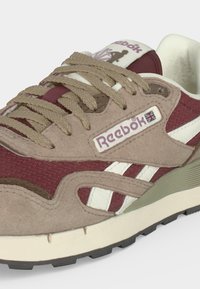Gros plan d'une basket Reebok beige et bordeaux avec des lacets beige, des accents blancs et un logo Reebok visible avec un drapeau Union Jack.