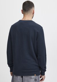 Mornarsko modra športna majica z dolgimi rokavi, okruglim ovratnikom, rebrastimi manšetami in robom. Ima majhno rjavo nalepko na spodnjem levem delu.