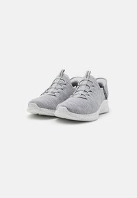 Des chaussures de sport grises avec une tige en tissu texturé, des lacets élastiques et une semelle blanche tachetée. Elles disposent d'une languette au talon pour un enfilage facile.