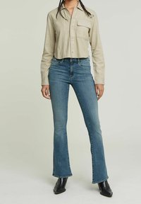 Ljust beige, kortärmad skjorta med knappar och bröstficka; blå utsvängda jeans; svarta klackstövlar; mjukt tyg och figurnära passform.