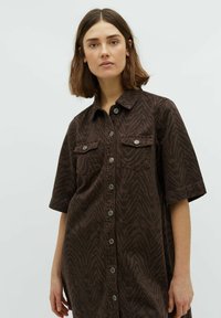 Frau mit schulterlangem braunem Haar, die ein braunes Knopfhemdkleid mit subtilm Zebramuster und Brusttaschen trägt.