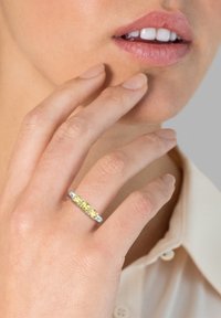 Silberring mit drei gelben Edelsteinen in einer linearen Anordnung, an einem Finger in der Nähe des Kinns getragen, mit natürlichen Hauttönen und einer leichten Bluse.