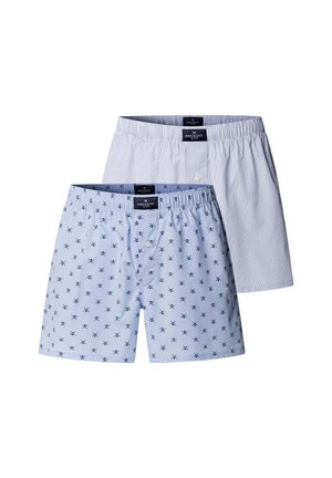 Jonge man met krullend haar draagt lichtblauwe boxershorts met een kleine donkere print, staat blootsvoets tegen een eenvoudige grijze achtergrond.