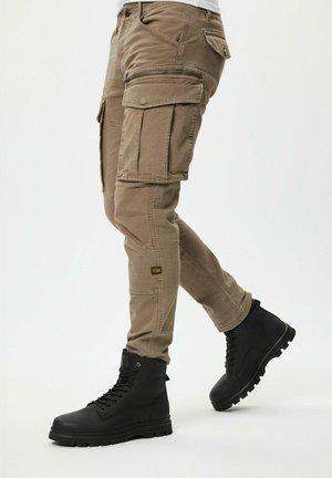 Osoba ubrana w beżowe spodnie typu cargo z wieloma kieszeniami i czarne buty na sznurówki, idąca na tle jednolitego białego tła.