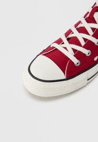 Κόκκινο πάνινο αθλητικό παπούτσι με λευκό λάστιχο στη μύτη και τη σόλα. Διαθέτει λευκά κορδόνια και μεταλλικά οπές με μαύρη γραμμή κοντά στη σόλα.