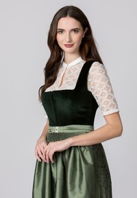 Grünes Samtkleid mit einem taillierten Oberteil und einem ausgestellten Rock, ergänzt durch eine weiße Spitzenbluse. Mit einem dekorativen Gürtel und floralen Akzenten.