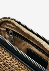 Borsa intrecciata in paglia color beige con rifiniture in pelle nera. Presenta una chiusura con zip che apre su un interno foderato in raso con una piccola tasca con zip.