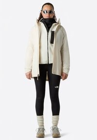 Giacca impermeabile beige chiaro, indossata sopra un pile nero con zip, abbinata a leggings neri, calzini crema al ginocchio e scarpe da trekking grigie.