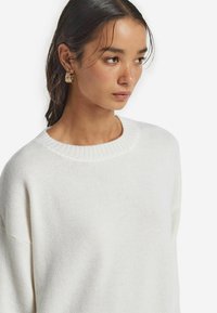 Weißer gestrickter Pullover mit rundem Ausschnitt, gerippten Bündchen und Oberteil. Der Stoff wirkt weich mit feiner Textur und minimalen Nähten.