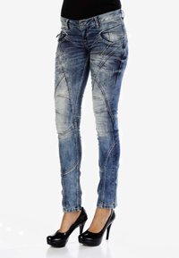 Jean skinny en denim bleu avec des accents déchirés, présentant des coutures en biais et une taille mi-haute. Associé à des talons hauts noirs.