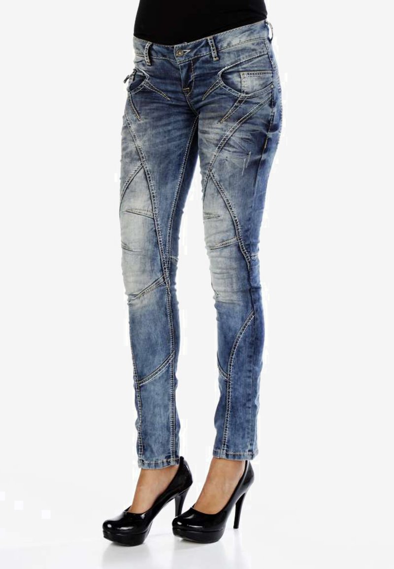 Jean skinny en denim bleu avec des accents déchirés, présentant des coutures en biais et une taille mi-haute. Associé à des talons hauts noirs.