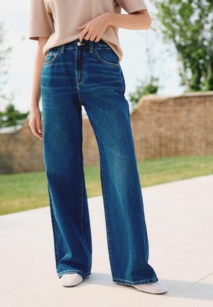 Person trägt weite blaue Jeans, beiges Kurzarmoberteil und weiße spitze Netzschuhe, steht draußen in der Nähe von Gras und einer Ziegelmauer.