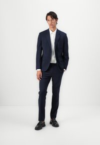Marineblauwe kostuum met een op maat gemaakte pasvorm, blazer met één knoop, bijpassende broek, wit overhemd en zwart leren instappers. Glad textuur van de stof.