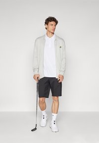 Light gray zip-up cardigan over a white polo, paired with black shorts. Holds a golf club, wearing white sneakers and crew socks.