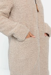 Beige Fleece-Mantel mit einer weichen, strukturierten Oberfläche, ausgestattet mit großen Taschen und einem Reißverschluss vorne. Die Ärmel und der Körper haben eine lässige Passform.