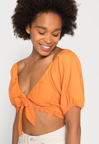 Top laranja curto com um laço à frente, mangas bufantes e bainha em babados. Textura suave e design em V, usado com calças de tom claro.