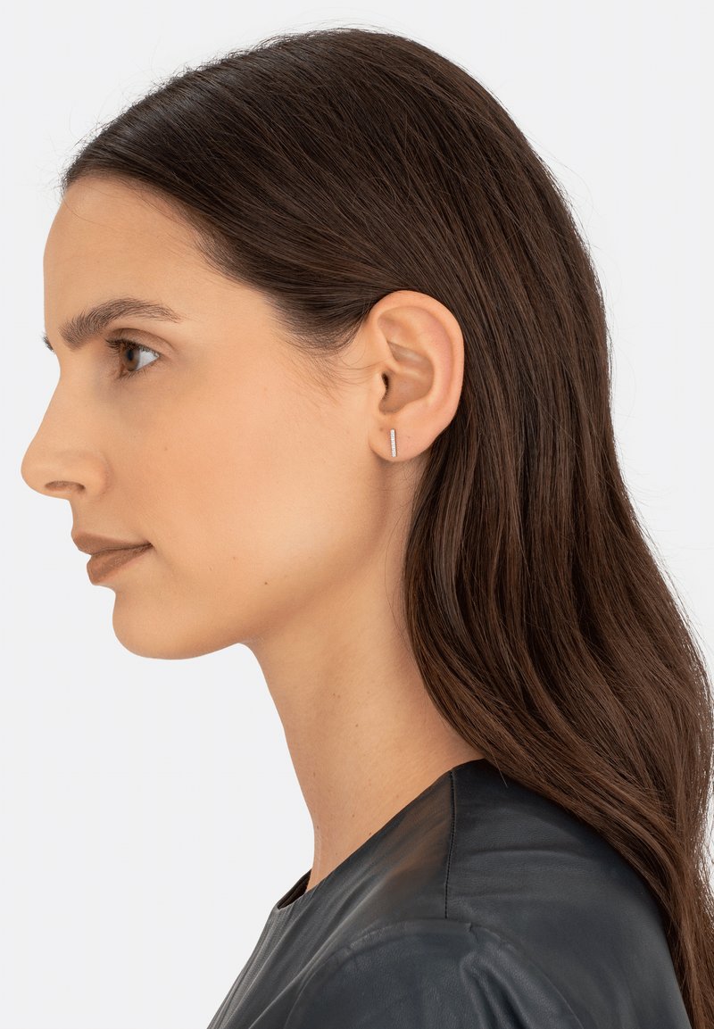 Silberne geometrische Steckerohrringe, die im Ohr eines Models getragen werden und ein schlankes, minimalistisches Design mit einer polierten Oberfläche präsentieren, das sich gegen dunkles Haar abhebt.