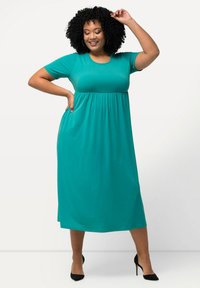 Vestido verde azulado de manga corta, hecho de una tela suave, con una cintura fruncida y bolsillos laterales, que llega hasta la mitad de la pantorrilla, combinado con zapatos de tacón negro.
