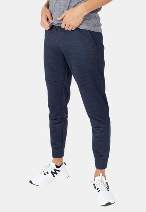 Personne portant un pantalon de jogging texturé bleu marine et des baskets blanches avec des détails noirs, les mains reposant près de la taille, debout devant un fond uni.