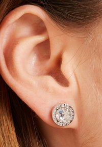 Zilveren stud oorbel met een heldere ronde kristallen centerpiece en de gravering "GUESS" rond de rand, weergegeven tegen een close-up van een oor.