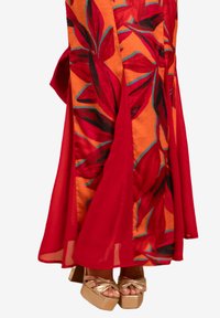 Lange rode jurk met bloemmotieven in tinten van rood en oranje, gecombineerd met gouden plateau-sandalen. Doorzichtige stof valt elegant.