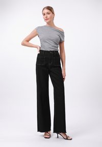 Graues Schulterfreies Oberteil kombiniert mit hochtaillierten, schwarzen, weiten Jeans mit kontrastierenden Nähten und zwei frontalen Taschen. Schwarze Riemchen-Heels.