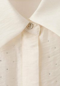 Cream-colored blouse with a pointed collar, button closure, and subtle embellishments of small rhinestones along the front.