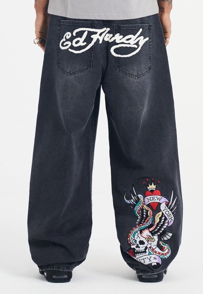 Leveled black denim-housut, joissa on valkoinen "Ed Hardy" -logo takana. Yksityiskohtainen brodeerattu graafinen kuva sydämestä ja pääkallosta kirkkailla väreillä.