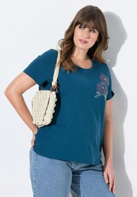 Turkoosi lyhythihainen t-paita, jossa on pinkkiä ja vihreää kukkaistikkaukset. Yhdistetty kermaiseen virkatusta olkalaukkuun, jossa on puinen hihna.