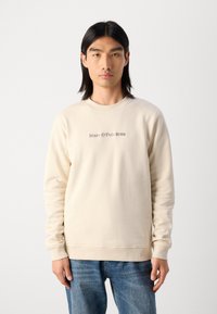 Šviesiai kreminis megztinis su raštuotu apykakle ir rankogaliais, kurio priekyje matomas subtilus išsiuvinėtas logotipas tamsesne siūle.