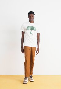 Hvid t-shirt med grøn "JORDAN" tryk og logo, parret med brune bukser og multicolor sneakers. Modellen står foran en ensfarvet baggrund.