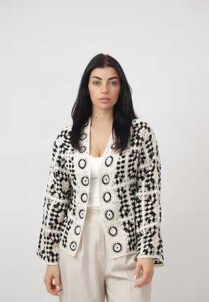 Kvinde med langt mørkt hår iført sort-hvidt mønstret hæklejakke over hvid top og lyse beige bukser, stående foran en ensfarvet baggrund.