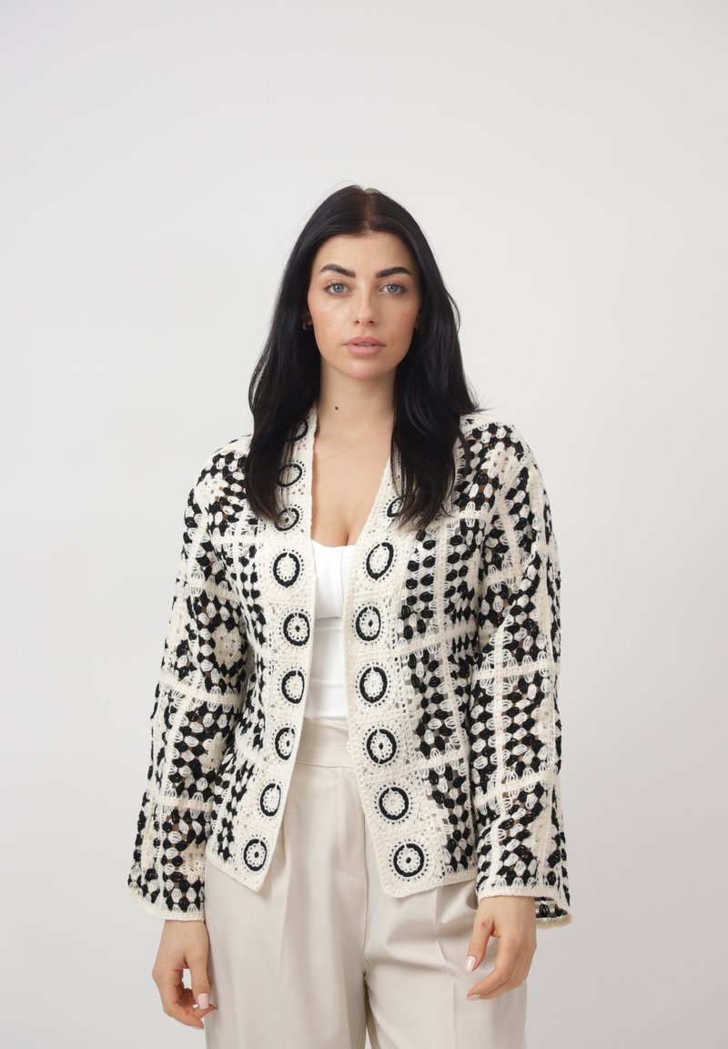 Mujer con cabello largo y oscuro, vistiendo una chaqueta de crochet con patrón en blanco y negro sobre una blusa blanca y pantalones beige claro, de pie frente a un fondo liso.