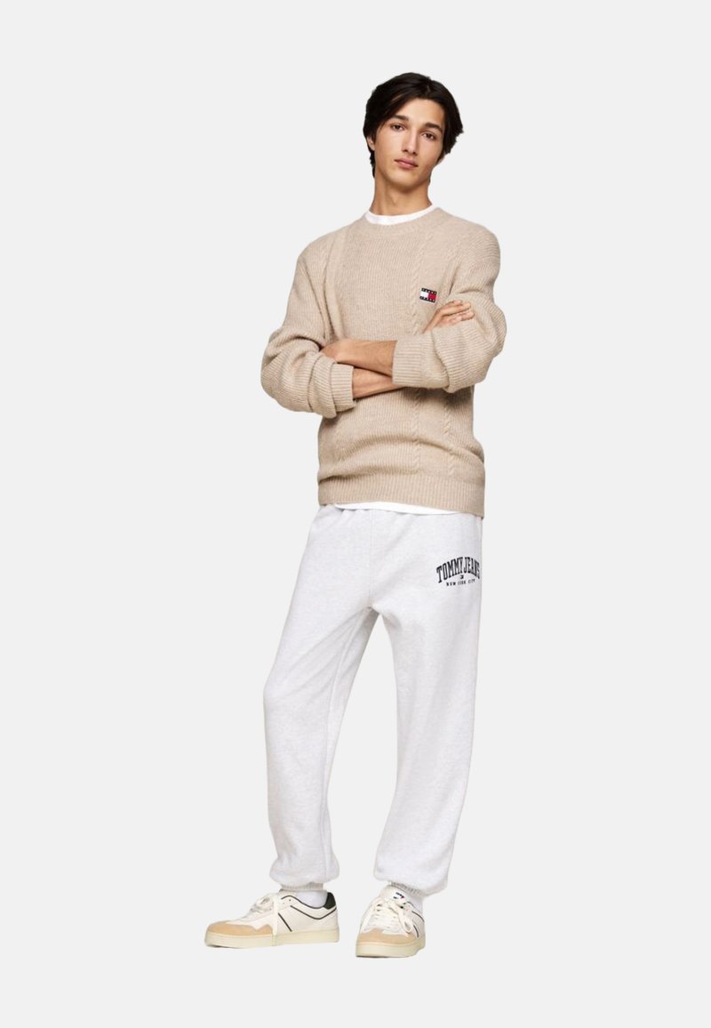 Joven de pie con los brazos cruzados, vistiendo un suéter beige, pantalones deportivos blancos con el texto "Tommy Jeans" y zapatillas beige, sobre un fondo blanco.
