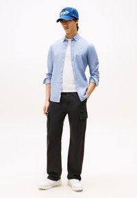 Svetlo modra srajca s sponkami, bela majica spodaj, črne cargo hlače z žepi in modra kapa, dopolnjena z belimi supergami.