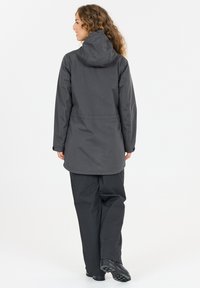 Frau mit lockigem Haar, die eine dunkelgraue Kapuzenjacke und passende Hosen trägt, steht mit dem Rücken zur Kamera vor einem weißen Hintergrund.