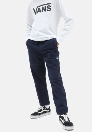 Granatowe dresowe spodnie z polaru z elastycznym paskiem, bocznymi kieszeniami i małym niebieskim logo Vans, w połączeniu z czarnymi butami do skateboardu i białą koszulą z długim rękawem.