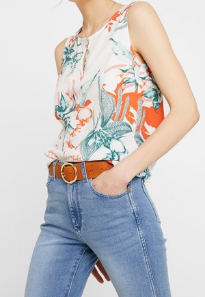 Femme portant un haut sans manches à motifs floraux avec des feuilles orange et vertes, rentrée dans un jean bleu clair, accessoirisée d'une ceinture marron et d'un collier en or.