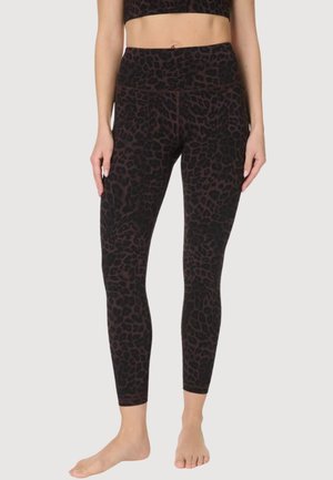 Vrouw draagt hoge taille, donkere luipaardprint legging en bijpassende sportbeha, staat blootsvoets tegen een effen achtergrond.