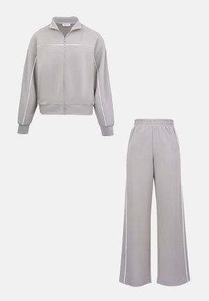 Giacca grigia chiaro con zip, rifinita con piping bianco, abbinata a pantaloni larghi con vita elastica e strisce laterali.