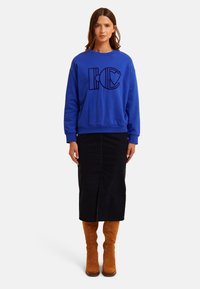 Blaues Sweatshirt mit schwarzen Grafikbuchstaben, kombiniert mit einem schwarzen Midirock und kniehohen, hellbraunen Stiefeln. Glatter Stoff, entspannter Schnitt, Rundhalsausschnitt.