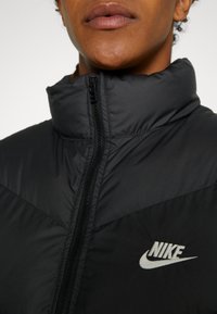 Chaqueta de plumas negra con cuello alto, cierre de cremallera y logo de Nike gris en el pecho. El material parece liso con un diseño acolchado.