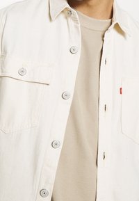 Camicia chiara con bottoni, due tasche sul petto, bottoni metallici e una morbida t-shirt beige sotto. Tessuto di cotone casual e testurizzato.